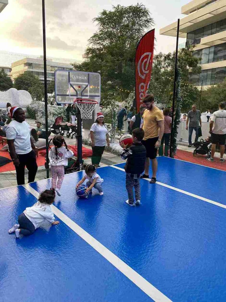 Adult helping a toddler shoot a basketball on a blue AirTrack mat at a UAE outdoor event.
